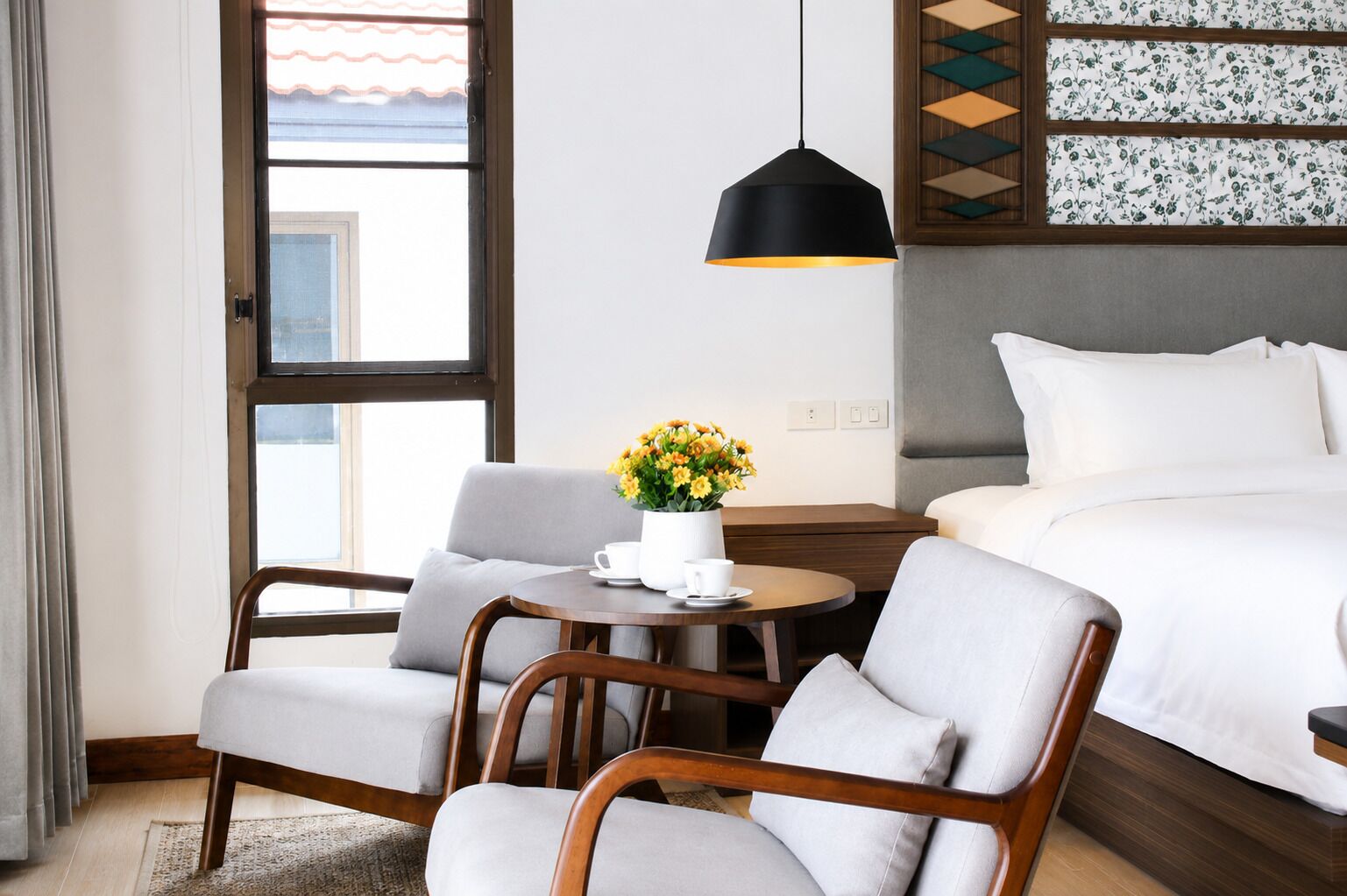 A hotel room with a bed, a desk with a chair, a coffee table with a flower vase, and a window with curtains.