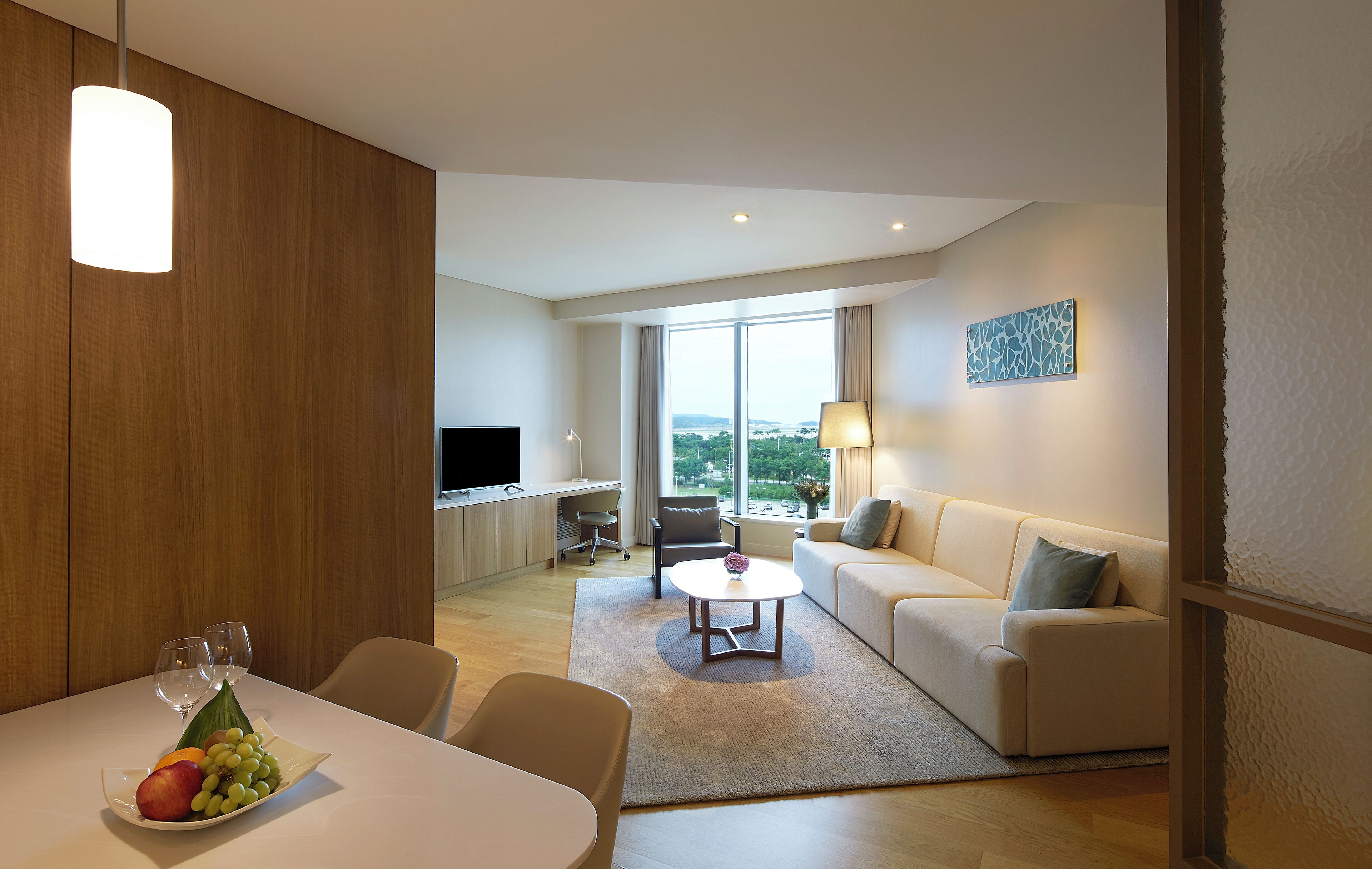 A modern hotel room with a dining area, a sofa, a TV, and a large window offering a city view.