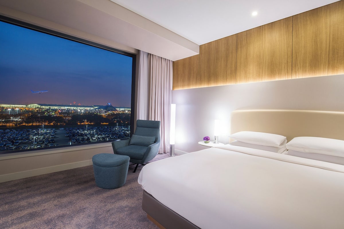 Una habitación de hotel con un ventanal grande, una cama, un sillón y una mesita de noche.
