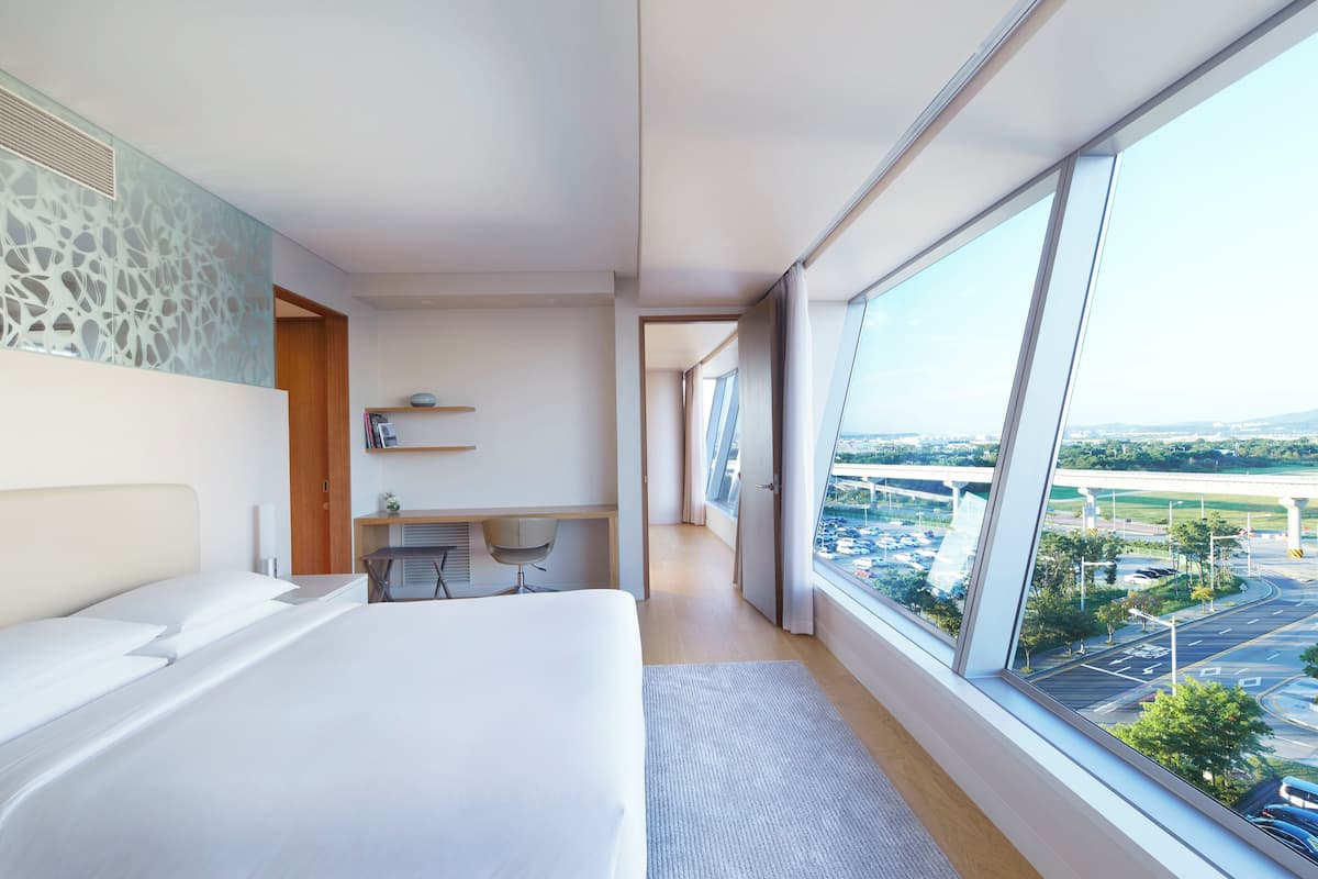 Habitación de hotel moderna con una cama grande, un escritorio y vistas a un paisaje urbano a través de amplias ventanas.