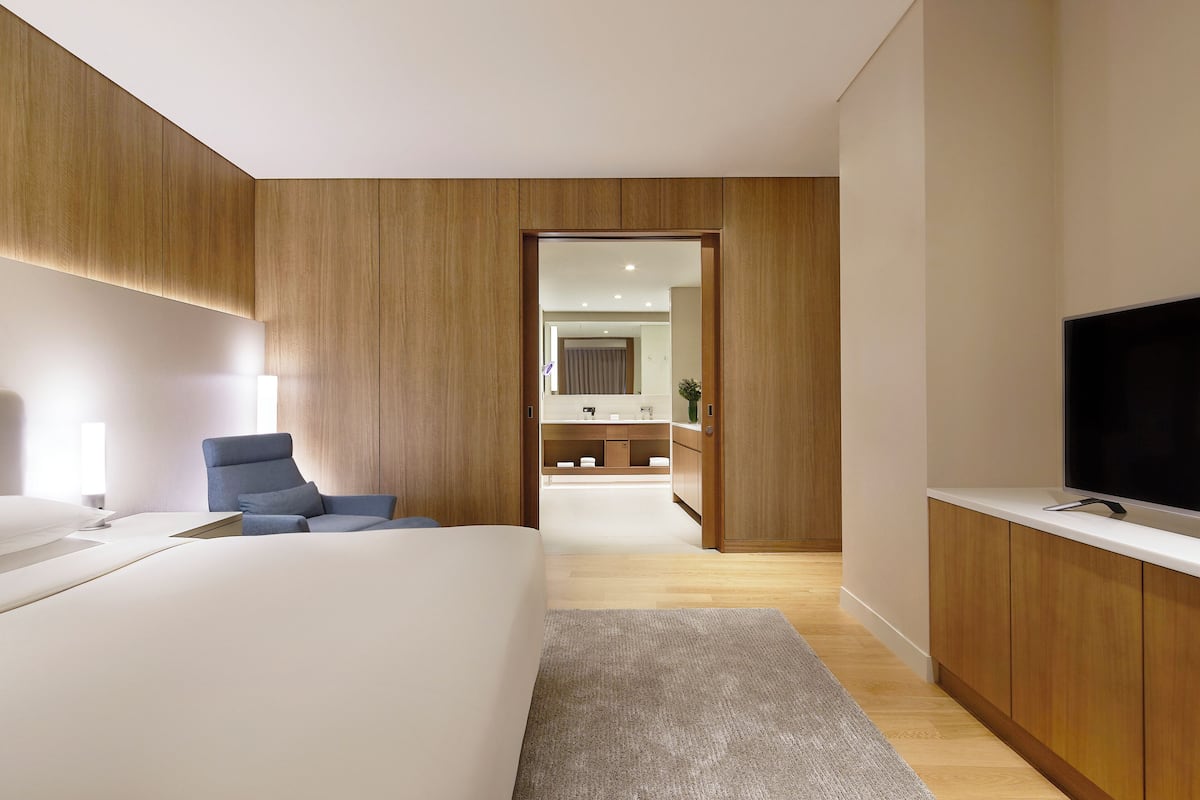 Una habitación de hotel moderna con una cama grande, una zona de estar con una silla azul, un televisor y paneles de madera en las paredes.