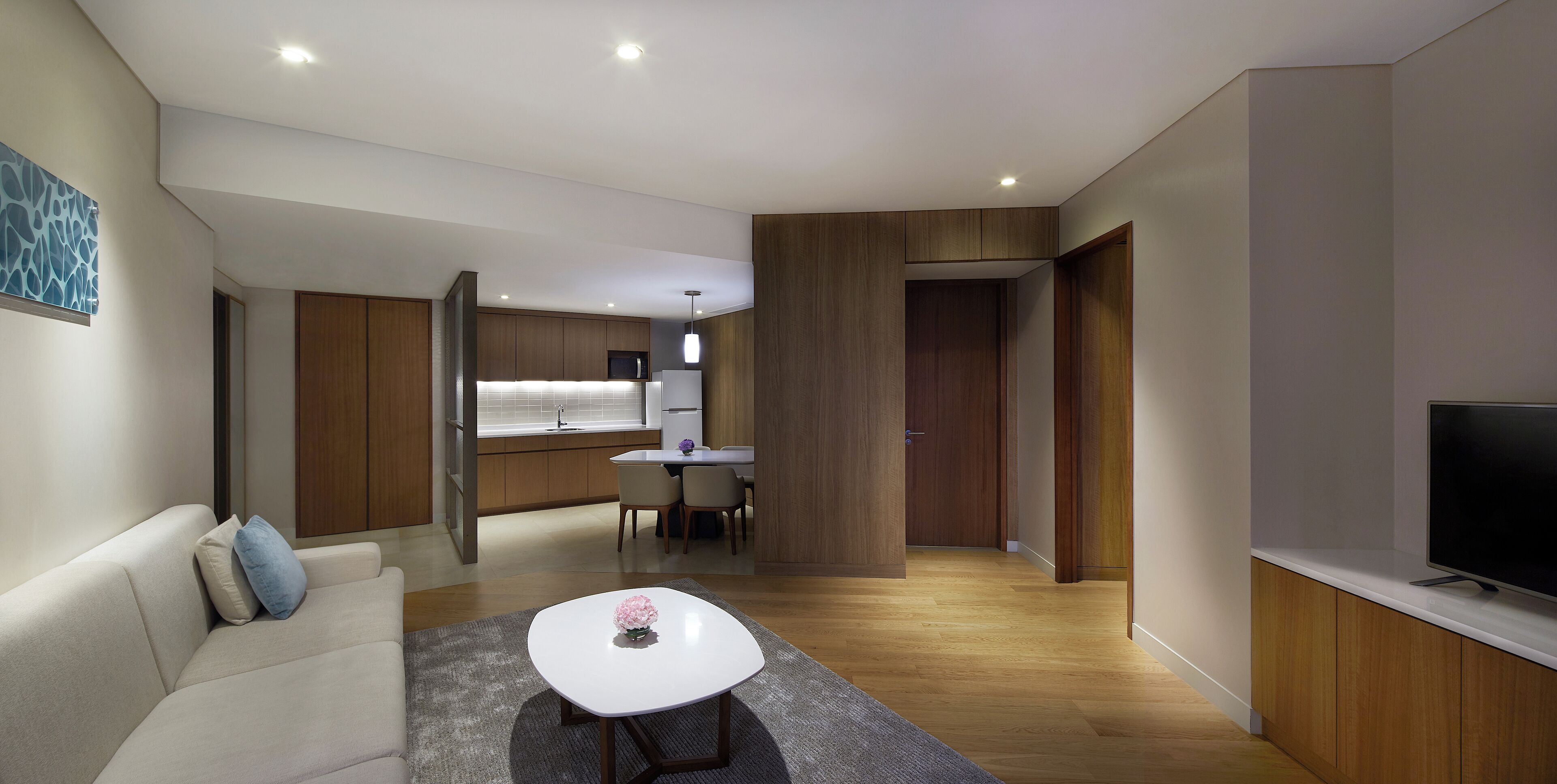 A modern living room with a sofa, a coffee table, and a TV. There is a dining area with a table and chairs, and a kitchen in the background.