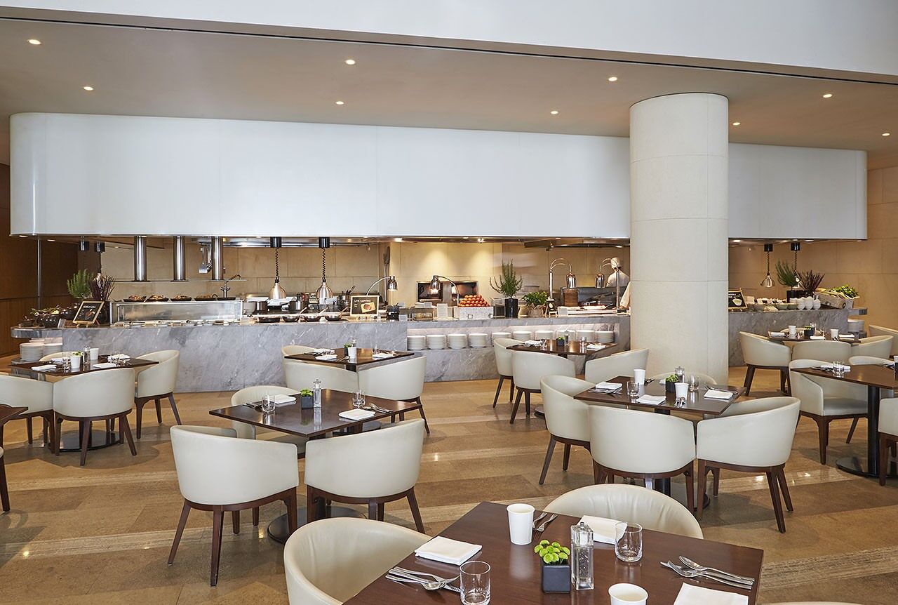 Een modern hotelrestaurant met meerdere tafels en stoelen, een centraal gelegen toonbank met diverse etenswaren, en een strak, minimalistisch design.