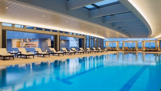 A modern indoor swimming pool with lounge chairs and a view of the cityscape through large windows.