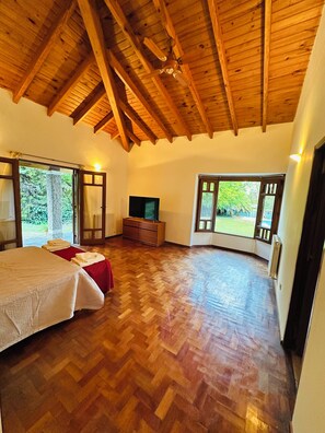 A spacious room with wooden flooring, a bed, a TV, and large windows offering a view of greenery. - Quinta los caracoles de chacras (quiet and comfortable) (Luján de Cuyo)