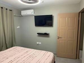 Ein Schlafzimmer mit einem Bett, einer Klimaanlage, einem Wand-Fernseher, einer Tür und einem an der Wand befestigten Regal.