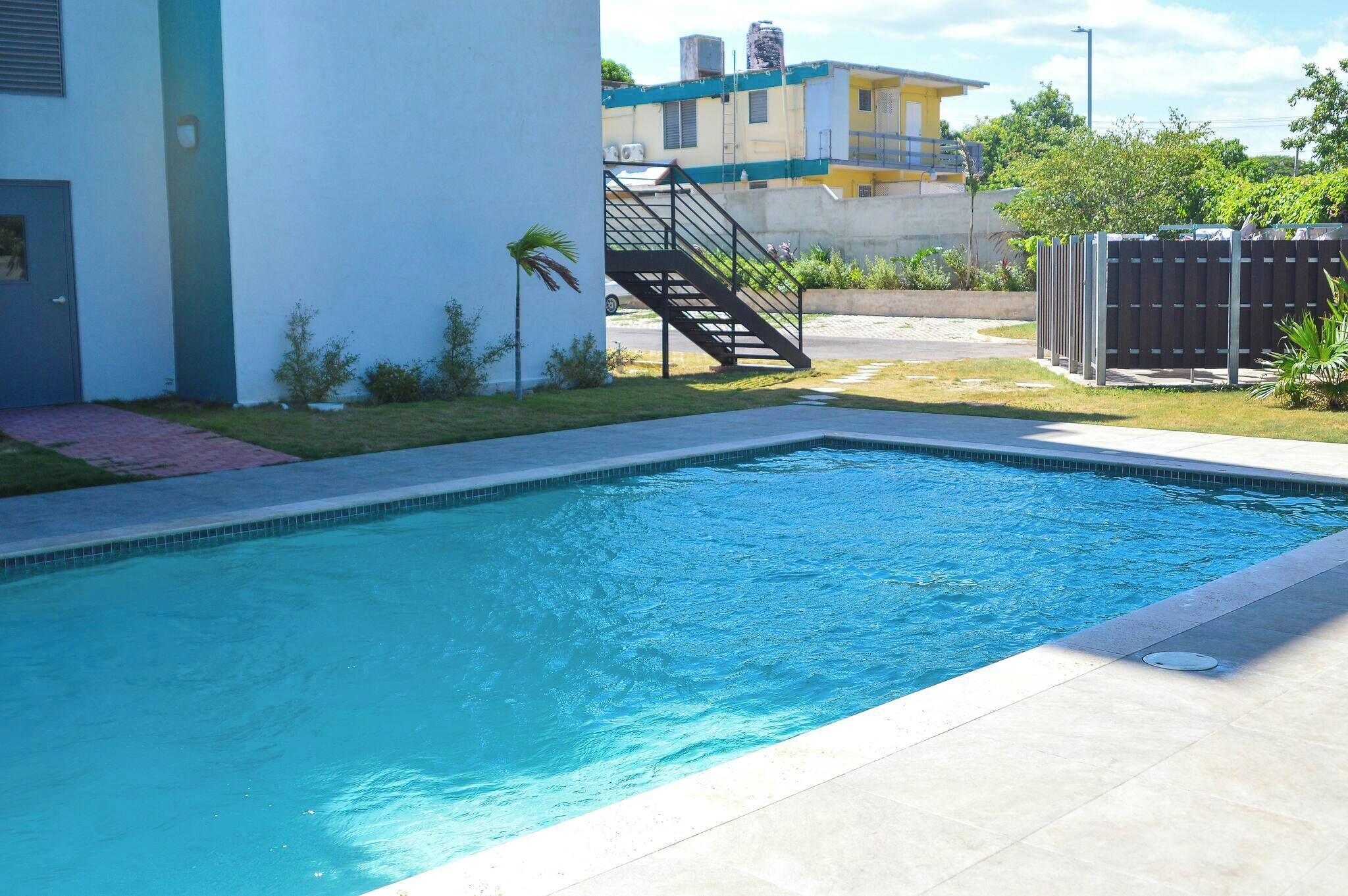A swimming pool with a tiled deck, a staircase leading to a building, and a fenced area with greenery.