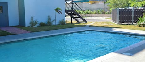 A swimming pool with a tiled deck, a staircase leading to a building, and a fenced area with greenery.