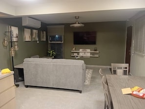 Ein Wohnzimmer mit grauem Sofa, einem Esstisch mit Stühlen, einem an der Wand montierten Fernseher und einem Deckenventilator.