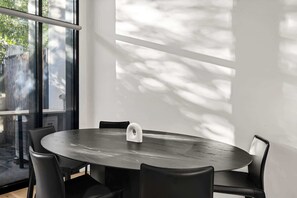 Mesa redonda com quatro cadeiras, um objeto branco sobre a mesa e vista para árvores através de grandes janelas.