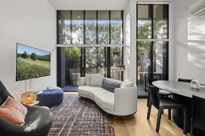 Una sala de estar moderna con un sofá blanco, una mesa de comedor con sillas, un televisor en la pared y ventanas amplias con vistas a zonas verdes.