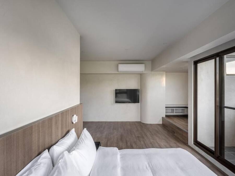 A modern hotel room with a bed, a flat-screen TV mounted on the wall, and a large window offering a view of the outdoors.