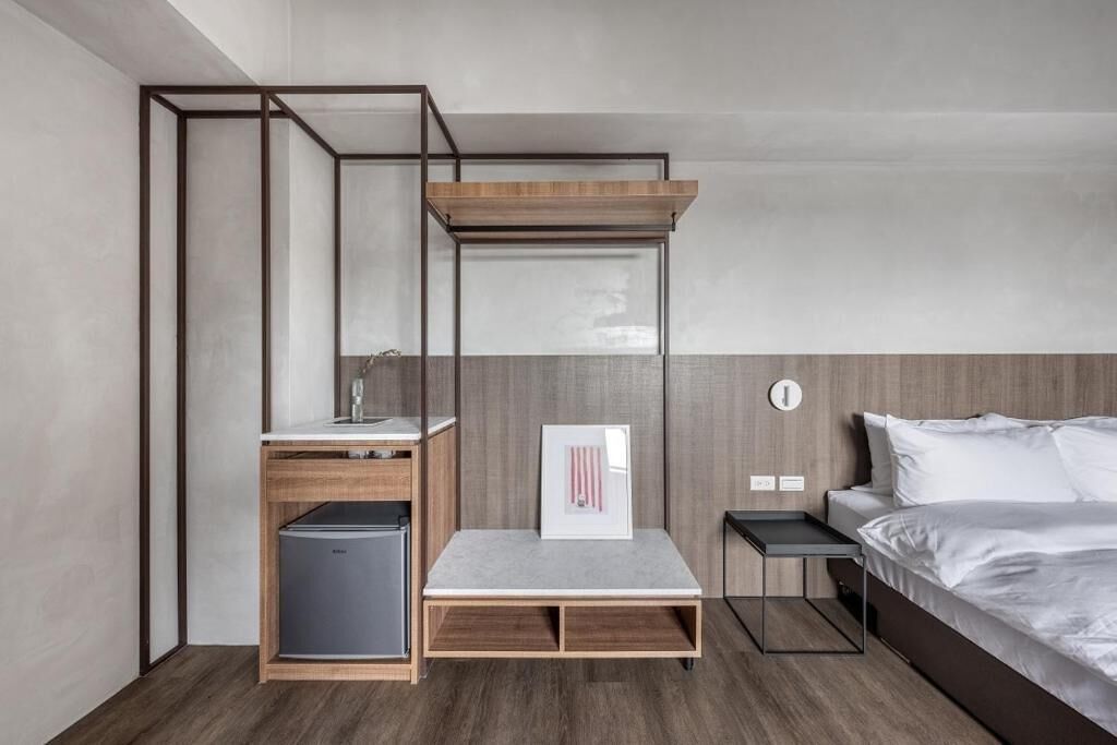 A compact hotel room with a bed, a small desk, a chair, a mini-fridge, and a built-in bench.