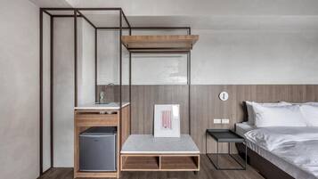 A compact hotel room with a bed, a small desk, a chair, a mini-fridge, and a built-in bench.