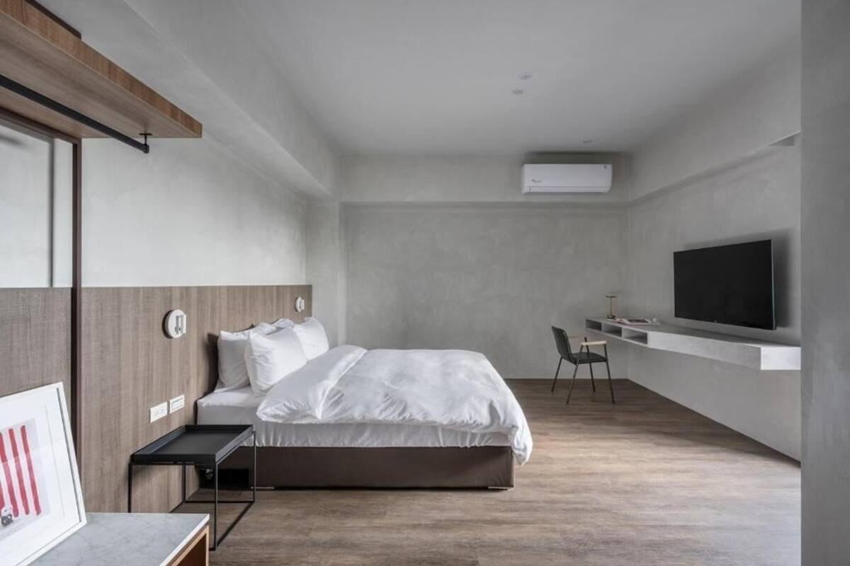 A modern hotel room with a large bed, a desk with a chair, a flat-screen TV mounted on the wall, and an air conditioning unit.