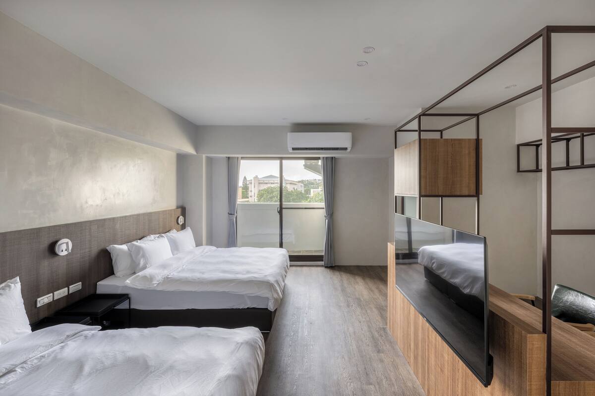 A modern hotel room with two beds, a large window, and a built-in TV.