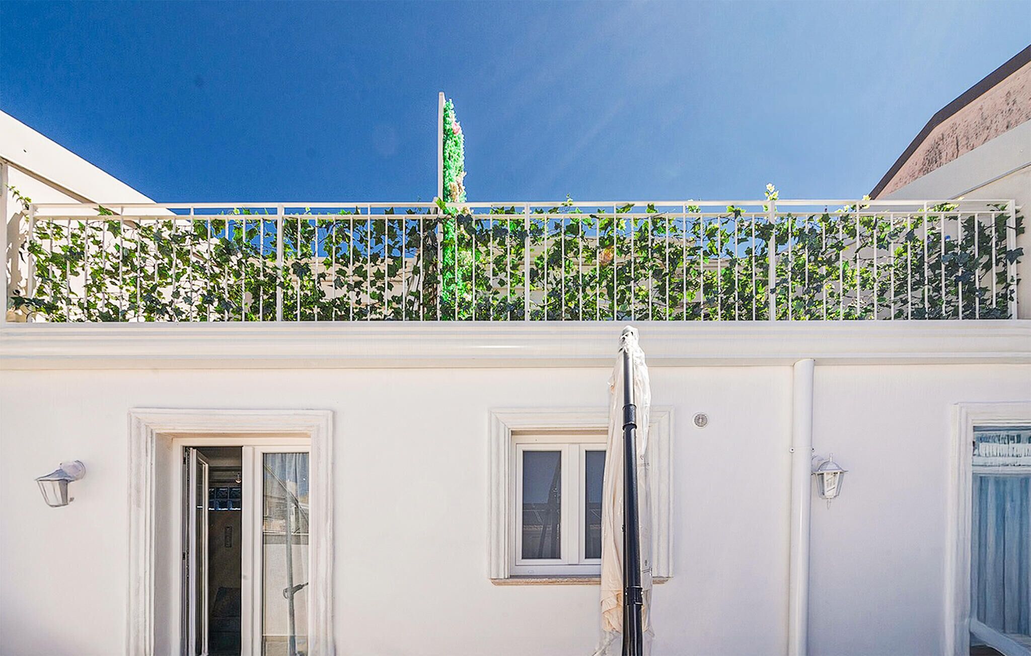 Un edificio con balcón cubierto de hiedra verde, paredes blancas y ventanas con cortinas.