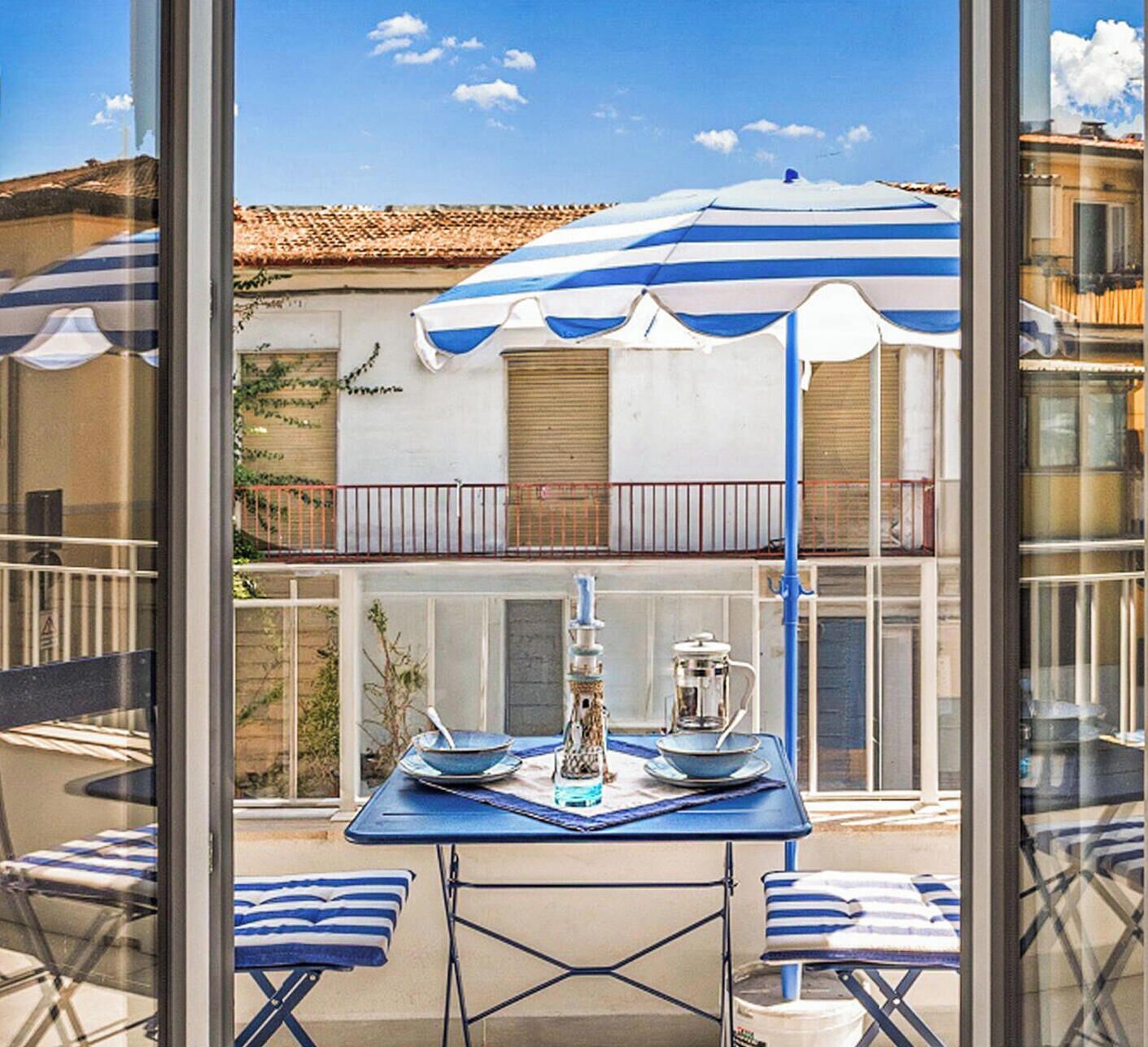 A balcony with a table set for two, a blue and white striped umbrella, and a view of a building with a tiled roof.