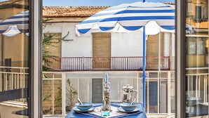 En balkon med et bord dækket til to, en blå og hvidstribet parasol og udsigt til en bygning med et tag af fliser.
