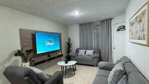 Una sala de estar con un sofá gris, un televisor montado en la pared y una mesita redonda con una planta.