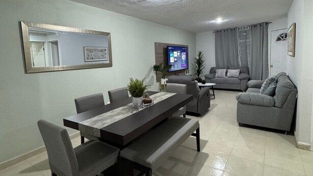 Un salón moderno con comedor, sofás grises, un televisor de pantalla plana y un espejo grande en la pared.