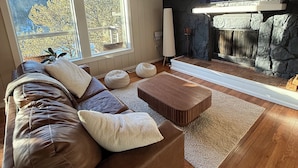 A cozy living room with a stone fireplace, wooden ceiling beams, and a large window overlooking a snowy landscape. - The Little House on the Hill - Families Look Here (Divide)