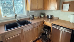 A modern kitchen with wooden cabinets, a stainless steel dishwasher, and a window providing a view of the outdoors. - The Little House on the Hill - Families Look Here (Divide)