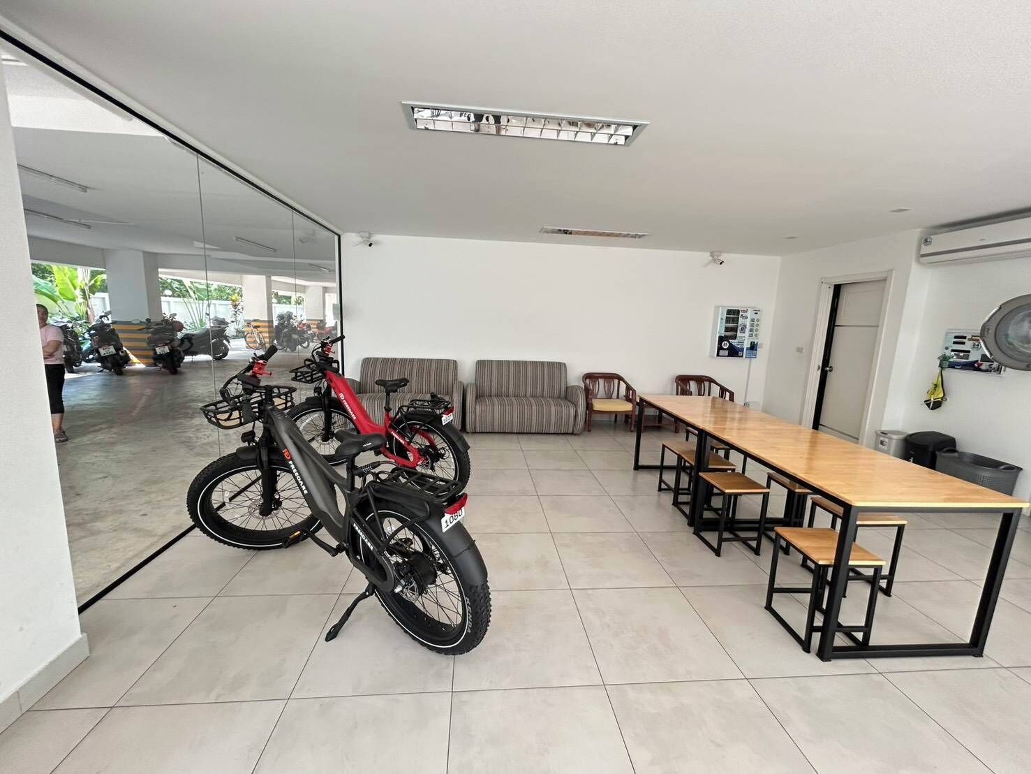 A spacious room with a long dining table, several chairs, a sofa, and a red electric bike parked near a glass door leading to a parking area with motorcycles.