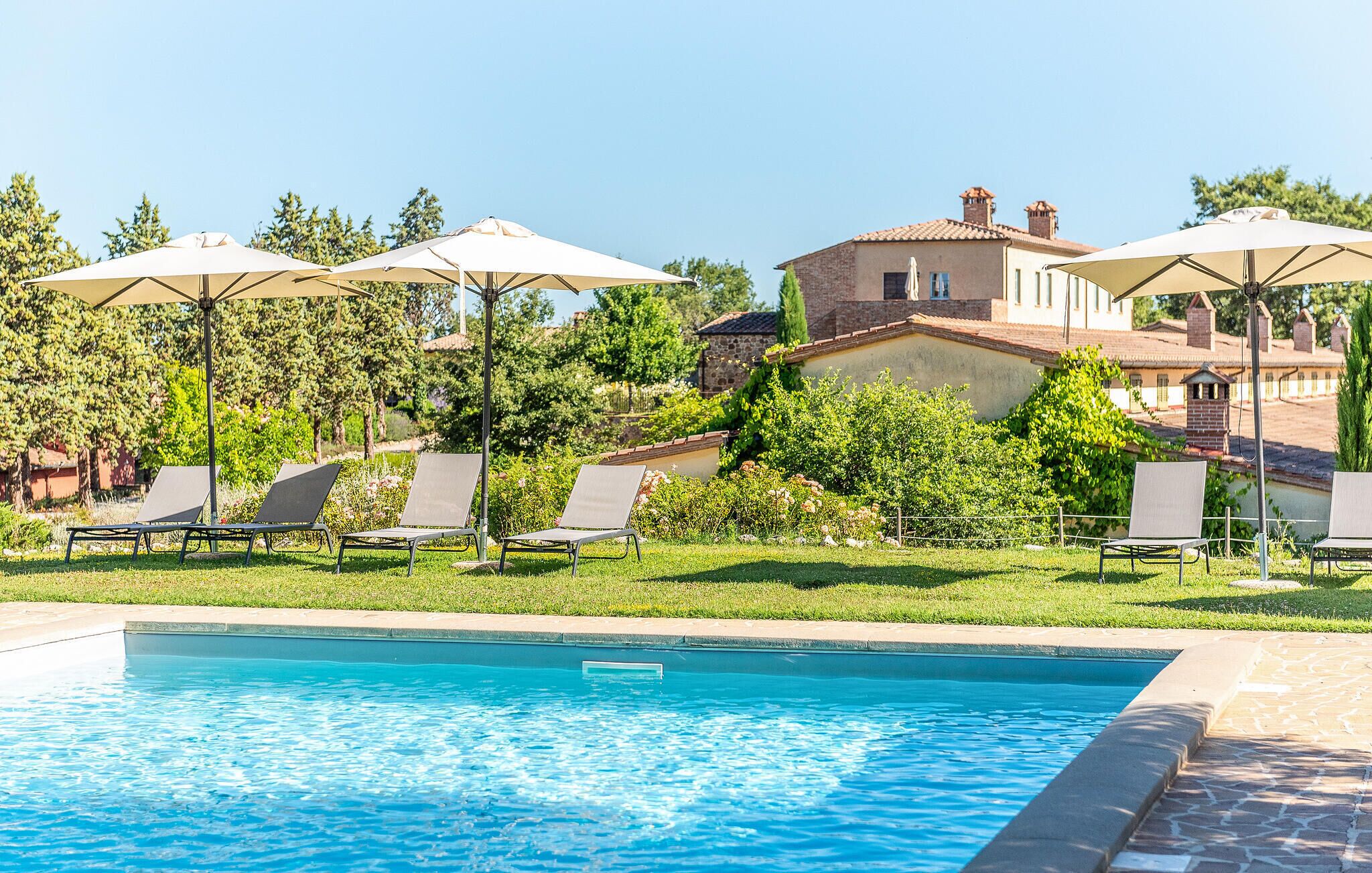 A swimming pool with lounge chairs and umbrellas, surrounded by greenery and a building with a tiled roof.