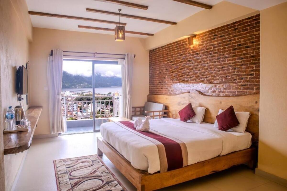 A bedroom with a large bed, a wooden headboard, a view of a cityscape through a glass door, and a small table with bottles.