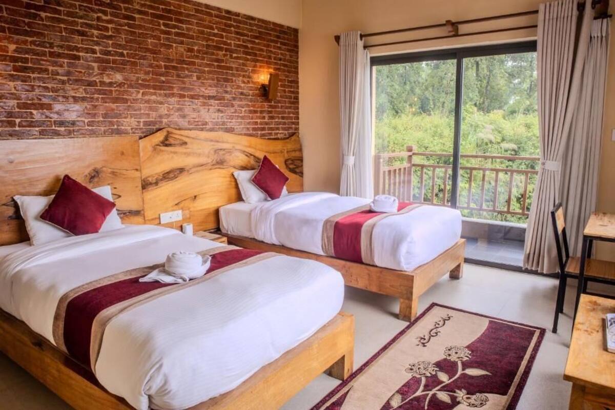 A hotel room with two wooden beds, a brick wall, a balcony with a view of greenery, and a table with a chair.