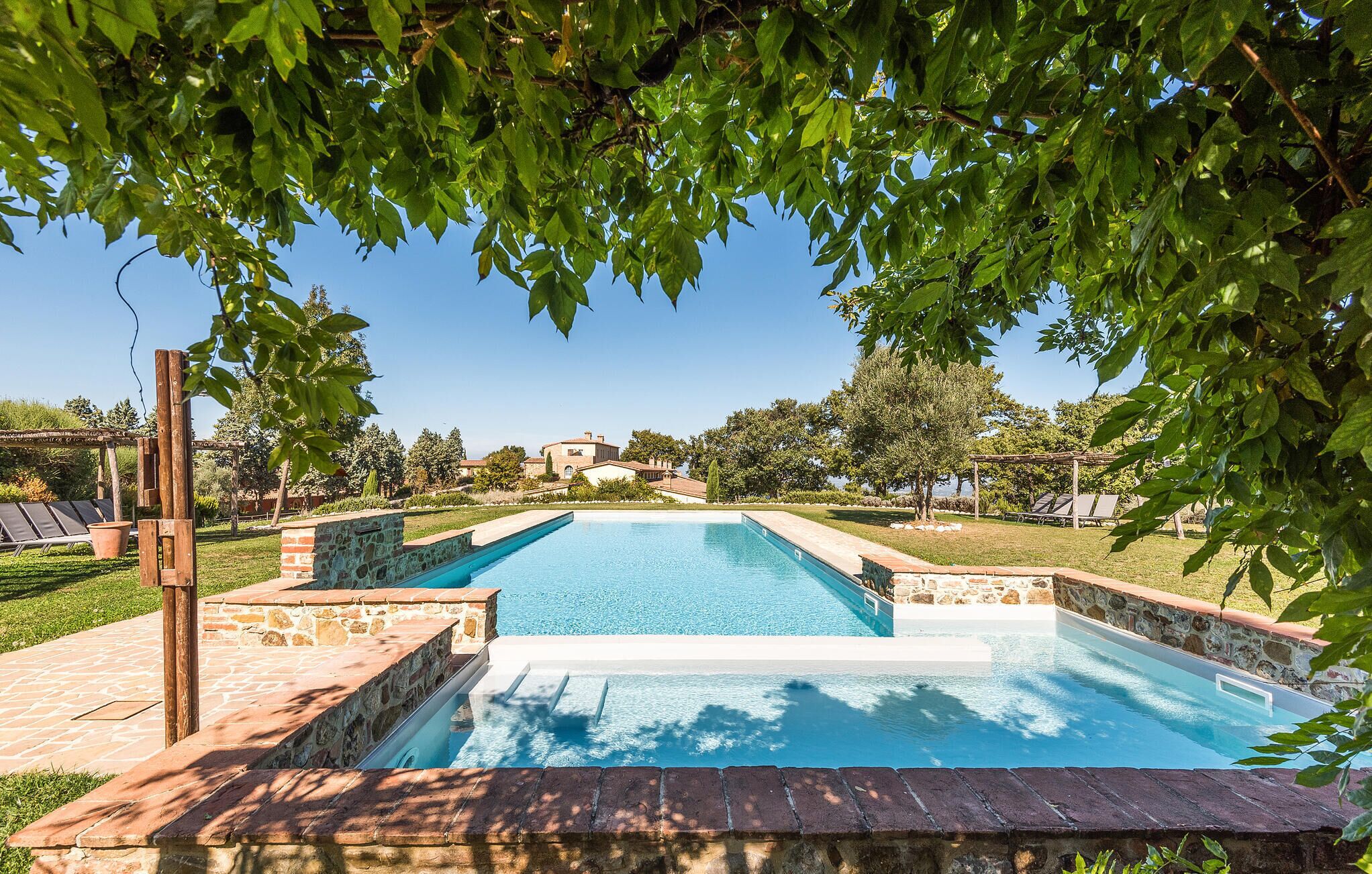 A rectangular swimming pool with stone and brick borders, surrounded by a grassy area and trees.