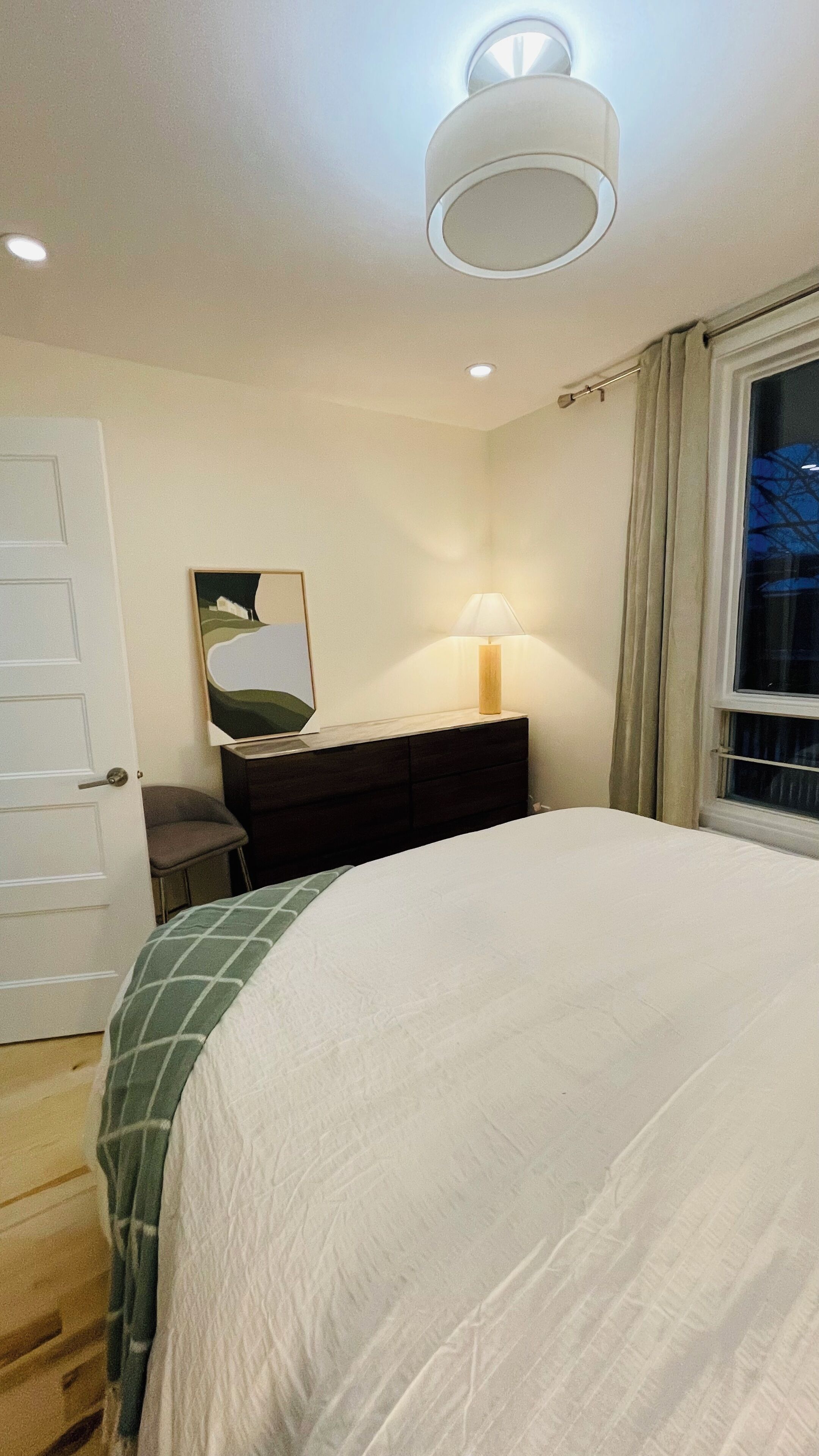 A bedroom with a bed, a bedside table with a lamp, a mirror, and a window with curtains.