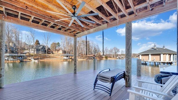 A wooden deck with a ceiling fan, overlooking a calm body of water with docked boats and houses in the background. - Serenity Shores - Private Lakefront Home with Dock (Moneta)
