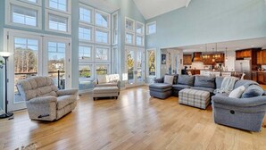 A spacious living room with a large sectional sofa, a recliner, and a coffee table. There are multiple large windows providing natural light and views of the outdoors. - Serenity Shores - Private Lakefront Home with Dock (Moneta)