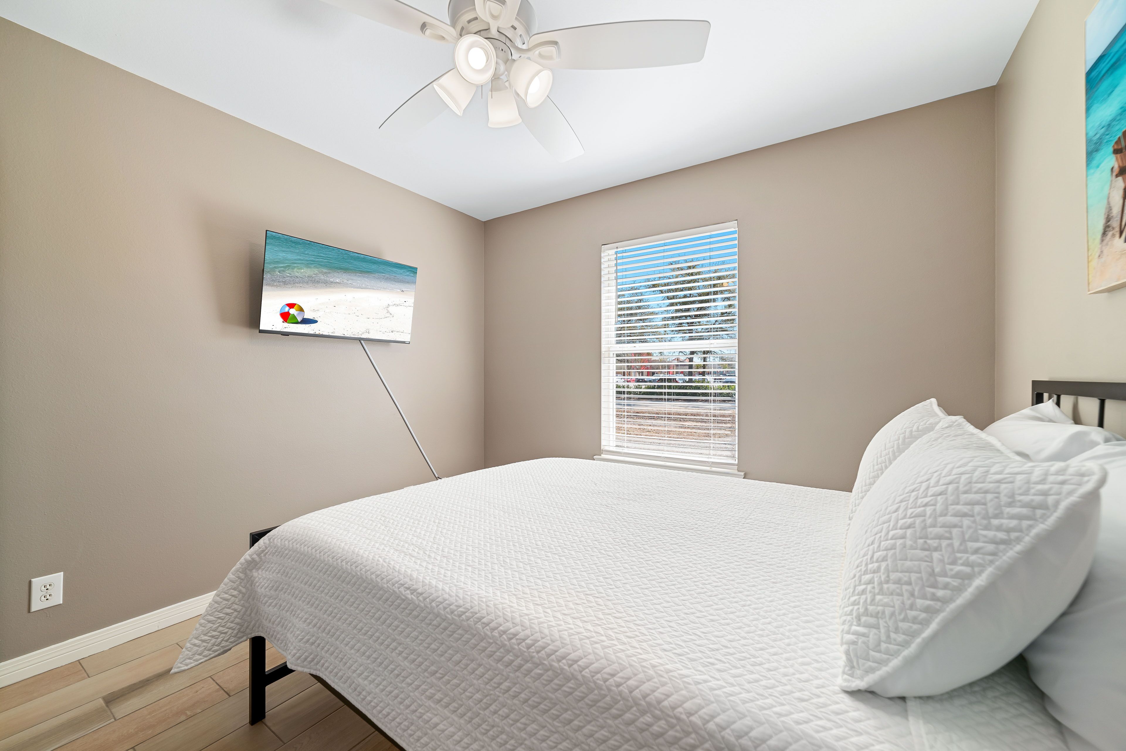 A bedroom with a bed, a ceiling fan, a wall-mounted TV, and a window with blinds.