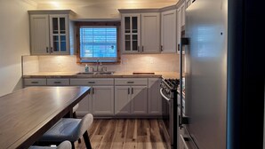 Eine moderne Küche mit weißen Schränken, einem Edelstahlkühlschrank, einem Holztisch mit Bänken und einem Fenster mit Jalousien.