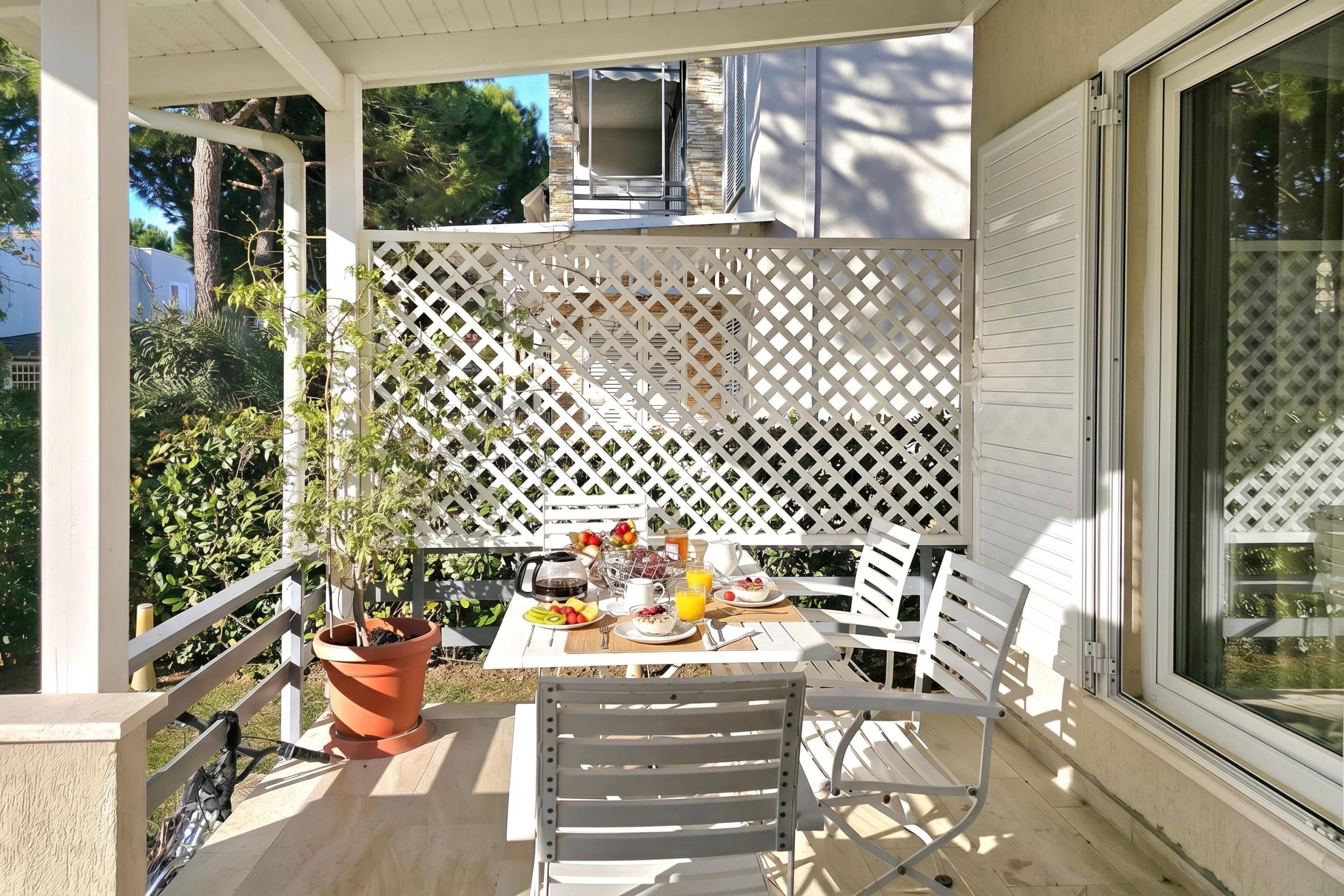 En overdækket terrasse med et bord dækket til morgenmad, en potteplante og en hvid stol.