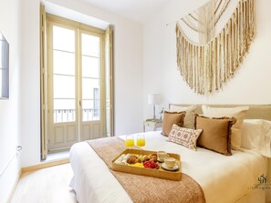 Une chambre d’hôtel avec un lit, un plateau de petit-déjeuner, une décoration murale en macramé et une fenêtre donnant sur des immeubles.