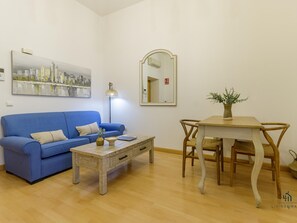 Ein Wohnzimmer mit blauem Sofa, Couchtisch aus Holz, Esstisch mit Stühlen, Stehlampe und Stadtbild an der Wand.