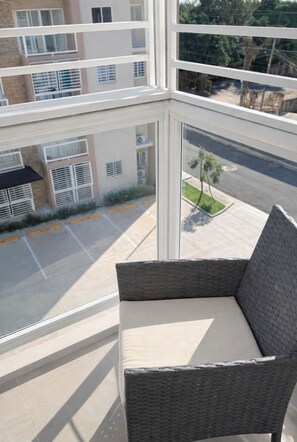 A balcony with a wicker chair, a view of a parking lot, and buildings in the background. - Modern 2-BR Apt. in Boca Chica l 5 min from the Airport 6 mins from beaches (Boca Chica)
