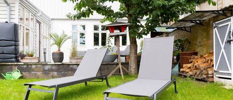 Two grey lounge chairs in a grassy backyard with a tree, a white building, and a stack of firewood.