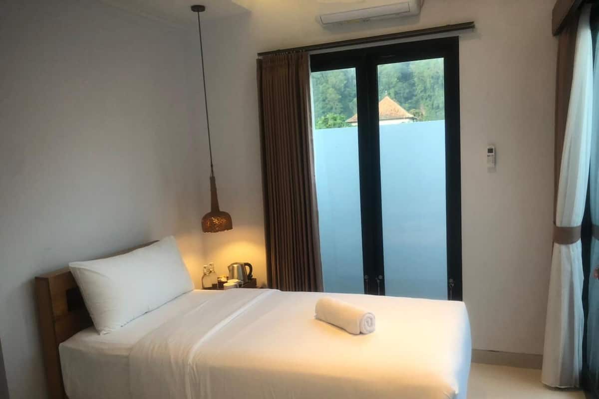 Habitación de hotel con cama, mesita de noche, lámpara, ventana con cortinas y vista a zonas verdes.