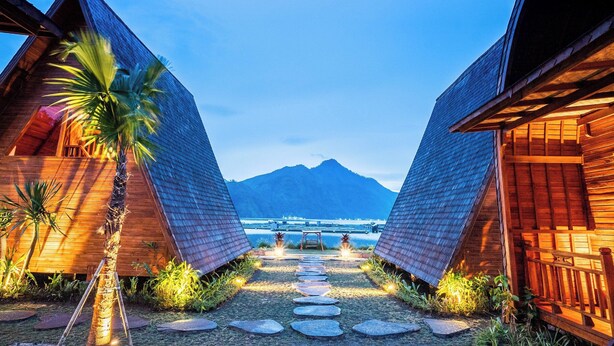 A pathway leading to two wooden cabins with a mountain and water body in the background. - The Dewi Tasik Kintamani - Cabin and Villa (Kintamani)