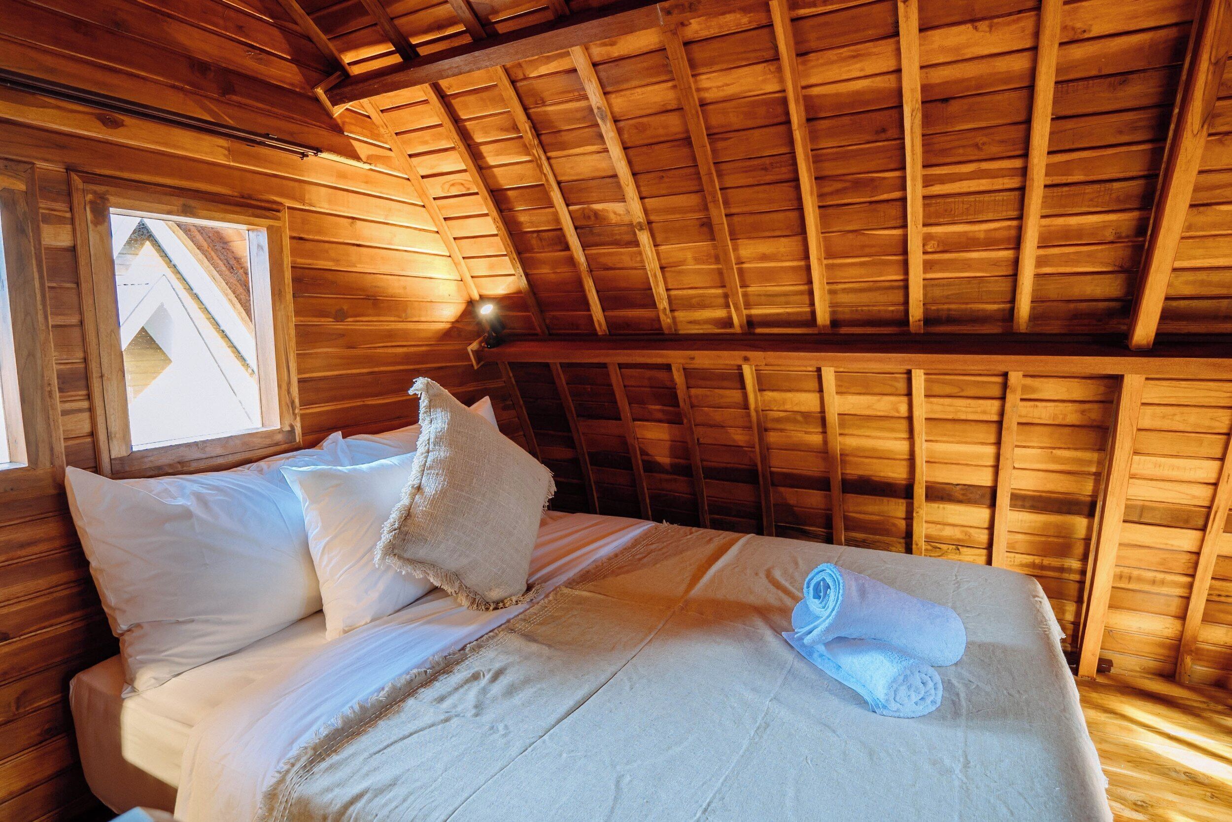A wooden room with a sloped ceiling, a bed with white linens, a window, and a lamp.