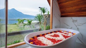 A bathtub filled with white and red flower petals, a wooden ceiling, and a view of a mountainous landscape through a window. - The Dewi Tasik Kintamani - Cabin and Villa (Kintamani)