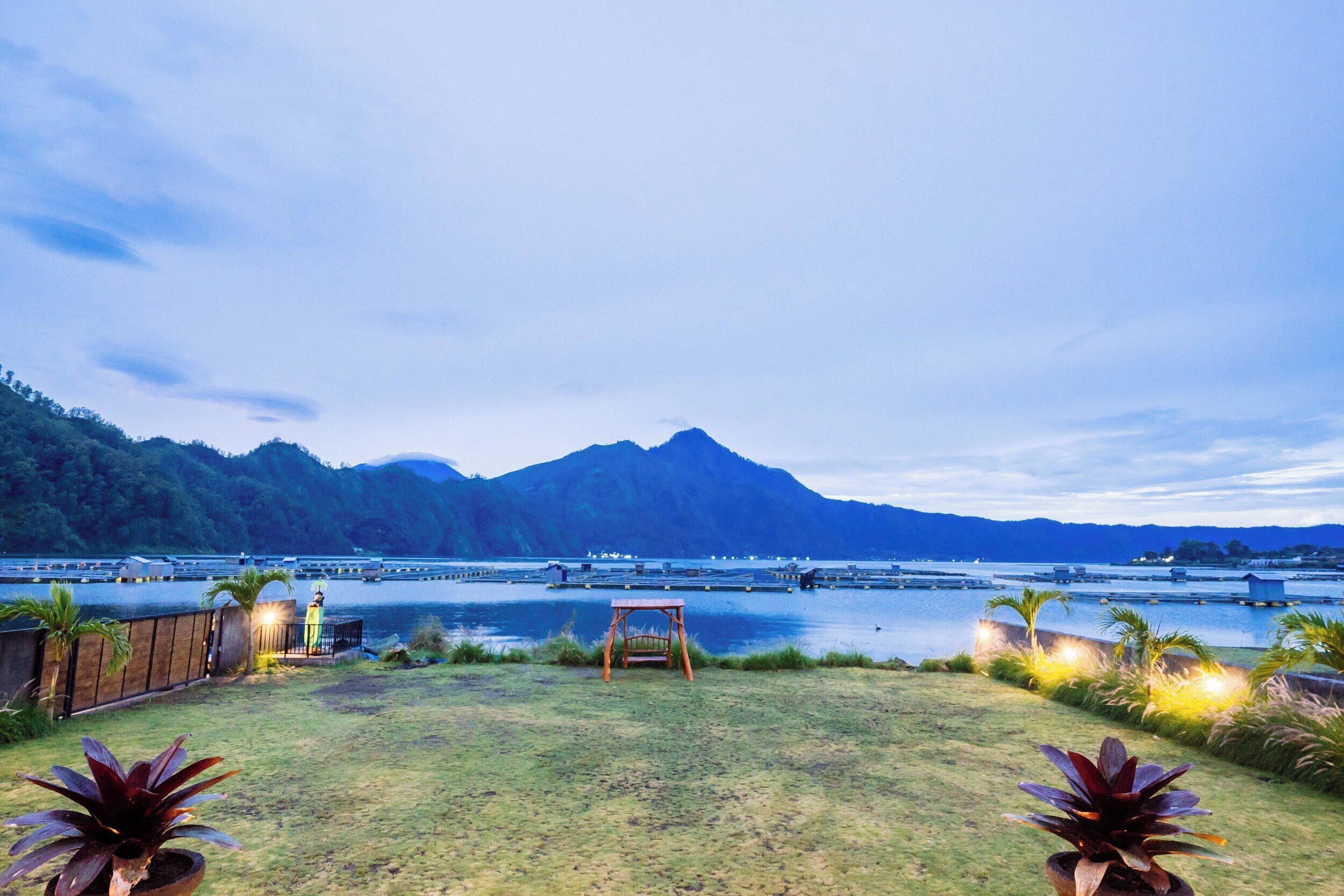 A grassy outdoor area with a swing set, a wooden fence, and a view of a body of water with mountains in the background.