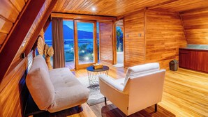 A cozy room with wooden walls and a sloped ceiling, featuring two sofas, a small round table, and a view of a mountain and water through large windows. - The Dewi Tasik Kintamani - Cabin and Villa (Kintamani)