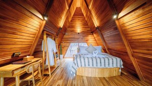 A cozy attic bedroom with wooden walls and a sloped ceiling, featuring a bed with a striped bedspread and two pillows, a wooden desk with a chair, and a small table with a lamp. - The Dewi Tasik Kintamani - Cabin and Villa (Kintamani)
