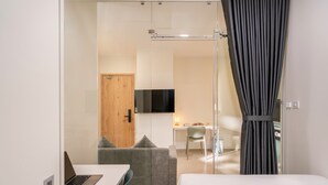 A modern hotel room with a bed, a desk with a laptop, a sofa, a TV mounted on the wall, and a glass door leading to a bathroom. - CityHouse NEST Bellita (Ho Chi Minh City)
